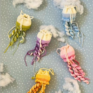 Op de afbeelding: Een verzameling handgehaakte kwallen in verschillende kleuren, waaronder geel, groen, paars, roze en blauw. Elke kwal heeft een ronde kop en lange, vloeiende tentakels. De achtergrond is een lichtblauwe, gestippelde ondergrond met witte wolken.