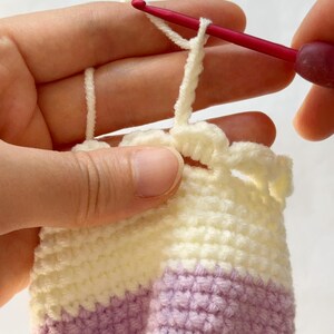 May include: Close-up of crochet work in progress. A red crochet hook is pulling a loop of cream-colored yarn through a stitch. The project features cream, lavender, and pale yellow yarn, creating a striped pattern. The work is held in a person's hands.