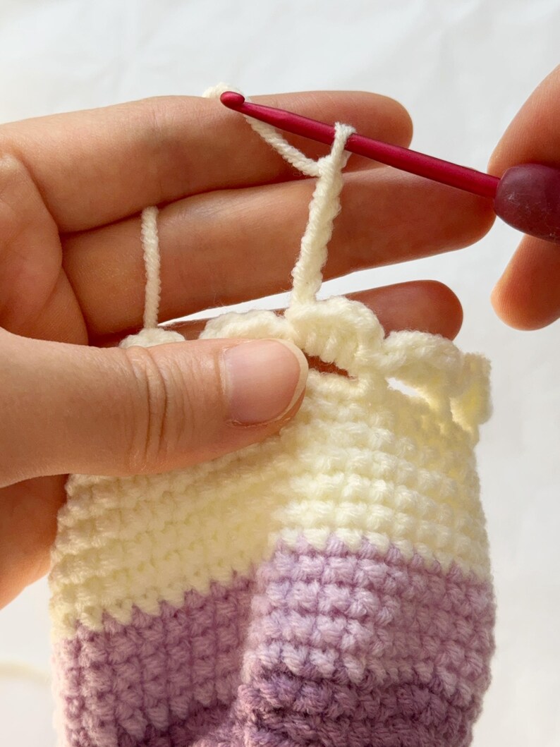 May include: Close-up of crochet work in progress. A red crochet hook is pulling a loop of cream-colored yarn through a stitch. The project features cream, lavender, and pale yellow yarn, creating a striped pattern. The work is held in a person's hands.