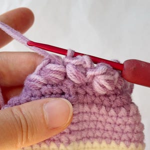 May include: Close-up of a crochet project in progress. The work features a gradient of colors, transitioning from yellow to lavender. A red crochet hook is inserted into the stitches, with a strand of lavender yarn attached.