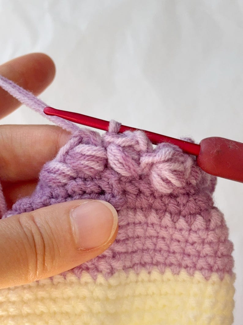 May include: Close-up of a crochet project in progress. The work features a gradient of colors, transitioning from yellow to lavender. A red crochet hook is inserted into the stitches, with a strand of lavender yarn attached.