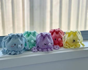 May include: A collection of five crocheted drawstring bags in pastel colors: light blue, mint green, lavender, coral, and yellow. Each bag features a cute octopus design with embroidered details and a ruffled base. The bags are displayed on a white surface.