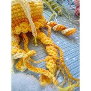 May include: A close-up of a handmade crochet jellyfish in shades of yellow and orange. The jellyfish has long, flowing tentacles and a flower-shaped charm. The background includes other crochet projects in various colors.
