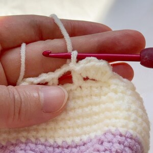 May include: Close-up of crochet work in progress. A hand holds a pink crochet hook, pulling white yarn through a loop. The project features alternating rows of white and lavender yarn, creating a textured fabric.