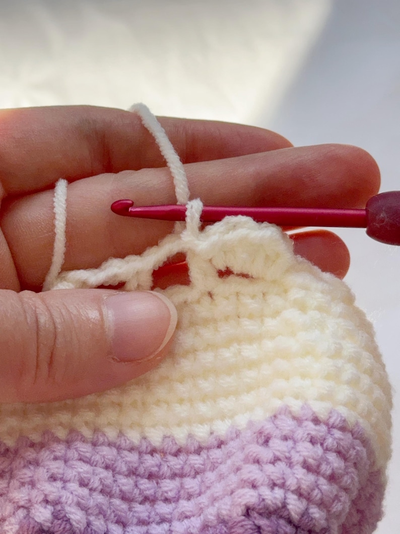 May include: Close-up of crochet work in progress. A hand holds a pink crochet hook, pulling white yarn through a loop. The project features alternating rows of white and lavender yarn, creating a textured fabric.