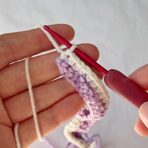 May include: Close-up of crochet work in progress. A pink crochet hook is inserted into a row of lavender and white yarn. The yarn is being worked into a curved shape. The background is a plain white surface.