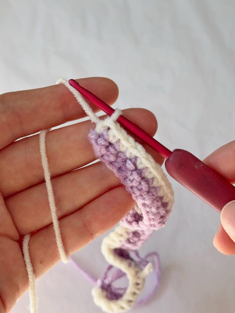 May include: Close-up of crochet work in progress. A pink crochet hook is inserted into a row of lavender and white yarn. The yarn is being worked into a curved shape. The background is a plain white surface.