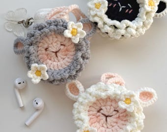 May include: Crocheted sheep-shaped AirPod cases in gray, white, and black, adorned with small white and yellow flowers. The cases have pink accents and are designed to hold wireless earbuds. Two white earbuds are also visible.