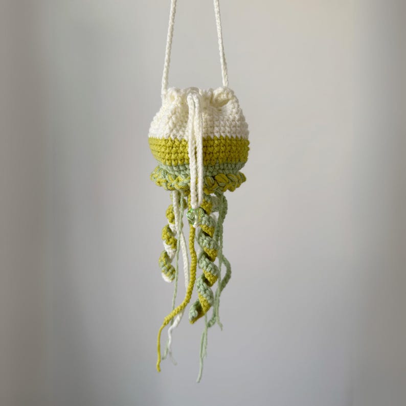 May include: A handmade crochet bag in the shape of a jellyfish. The bag is white with a yellow-green band and a ruffled bottom. Long, spiraling tentacles in shades of green and yellow dangle from the bottom, and a white strap is attached.