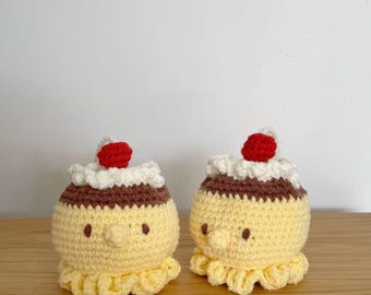 May include: Two handmade crochet dessert toys. Each has a yellow body, a brown band, white frosting, and a red cherry on top. The toys have small brown eyes and a ruffled yellow base. They are sitting on a wooden surface.
