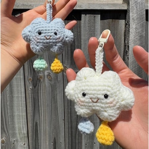 May include: Two handmade crochet cloud keyrings. One is light blue, the other is white. Each cloud has a smiling face, pink cheeks, and colourful rain drops. The keyrings are held up by a white strap and a silver clasp.