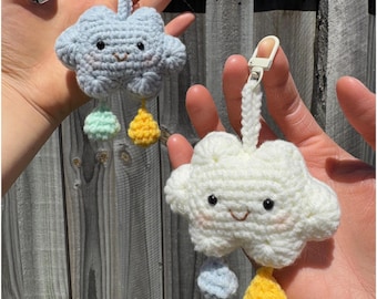 May include: Two handmade crochet cloud keychains. One is light blue, the other is white. Each cloud has a smiling face, pink cheeks, and colorful rain drops. The keychains are held up by a white strap and a silver clasp.