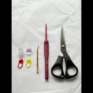 May include: A collection of crochet tools, including a burgundy crochet hook, a yarn needle, stitch markers, clear and purple stitch holders, and black-handled scissors. The crochet hook is labeled "5/0 3.0mm".