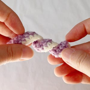 May include: A close-up of a crocheted item held between two hands. The item is a twisted rope design, with alternating sections of lavender and white yarn. The background is a soft, neutral color.