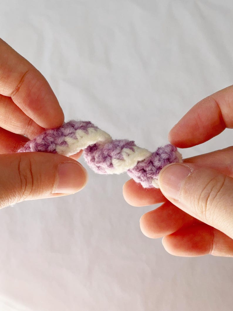 May include: A close-up of a crocheted item held between two hands. The item is a twisted rope design, with alternating sections of lavender and white yarn. The background is a soft, neutral color.