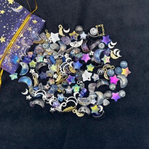 May include: A selection of celestial charms and beads, featuring moons, stars, and feathers in silver, gold, blue, purple, and iridescent colours. A small purple pouch with gold detailing is visible. The items are scattered on a dark surface.
