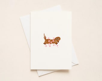 Könnte beinhalten: Eine weiße Grußkarte mit einer braunen Dackel-Illustration und rosa Herz-Akzenten. Die Karte wird von einem weißen Umschlag begleitet, perfekt zum Versenden einer herzlichen Nachricht. Das Design ist schlicht und charmant.