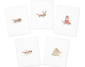 Könnte beinhalten: Set mit fünf weißen Grußkarten mit Aquarellillustrationen von Dackeln in festlichen Urlaubsszenen. Jede Karte zeigt ein anderes Dackel-Design, darunter eines mit Geweih und ein weiteres mit einem Weihnachtsbaum.