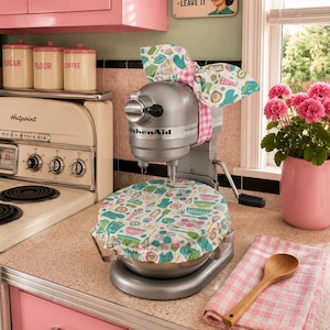 May include: A silver KitchenAid stand mixer adorned with a fabric cover featuring a colorful kitchen-themed print of bowls, whisks, and spatulas. A matching fabric bow is tied around the head of the mixer with a pink and white gingham ribbon.