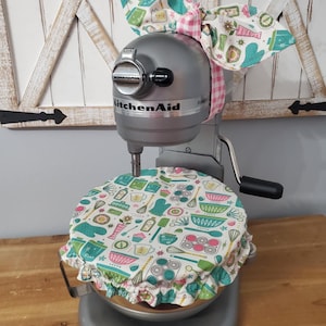 May include: A silver KitchenAid stand mixer with a patterned bowl cover and a matching bow. The cover and bow feature a white background with teal, pink, and yellow kitchen-themed illustrations. The mixer is on a wooden surface.