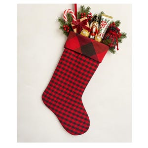 May include: A red and black buffalo check Christmas stocking filled with a candy cane, a small gift box with a velvet ribbon, a nutcracker figurine, two gold-wrapped chocolates, a tube of cream, and a small plaid drawstring pouch.