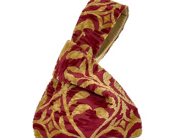 May include: A wristlet bag featuring a deep red and gold woven fabric with a repeating leaf and geometric pattern. The bag has a unique knot-style handle where one side is longer than the other, creating a distinct asymmetrical silhouette.