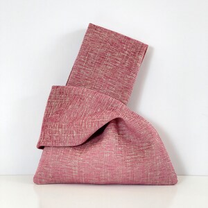 May include: A pink fabric tote bag with a knotted design. The bag is made of a textured fabric with a subtle white pattern.