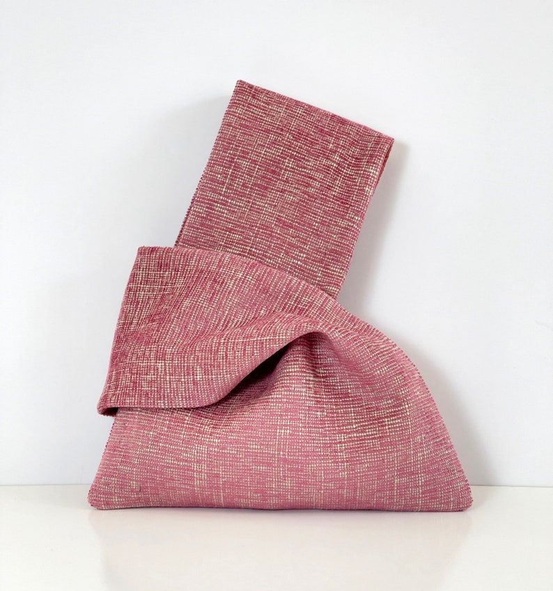 May include: A pink fabric tote bag with a knotted design. The bag is made of a textured fabric with a subtle white pattern.