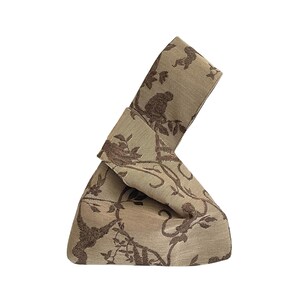 May include: A beige fabric handbag with a brown botanical pattern featuring monkeys and foliage. The bag is folded and knotted, creating a unique, structured shape. The fabric has a woven texture.