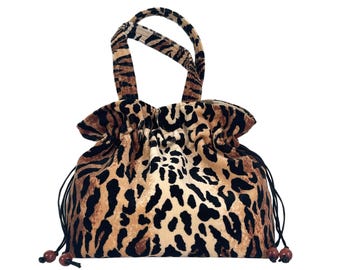 May include: A drawstring handbag featuring a tan and black leopard print pattern on a soft-textured fabric. The bag includes two matching top handles, a gathered top opening, and black cord drawstrings finished with small, round, dark brown beads.