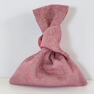 May include: A pink fabric bag with a knotted handle. The bag is made of a textured fabric with a subtle pattern.