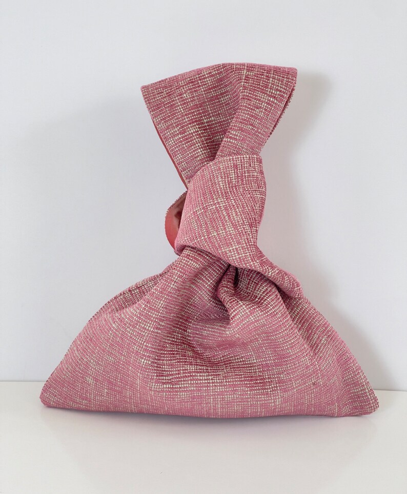May include: A pink fabric bag with a knotted handle. The bag is made of a textured fabric with a subtle pattern.