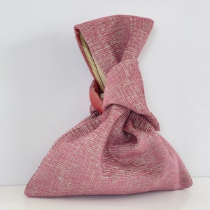 May include: A pink fabric bag with a knotted handle. The bag is made of a textured fabric with a subtle pattern.