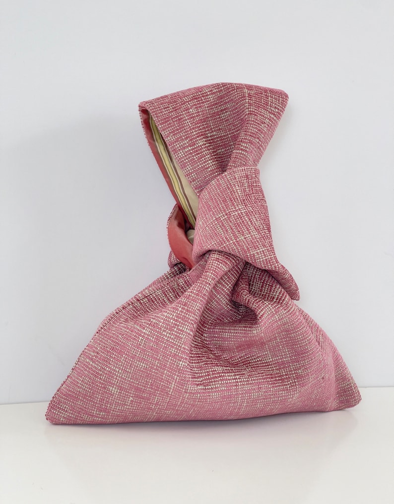 May include: A pink fabric bag with a knotted handle. The bag is made of a textured fabric with a subtle pattern.