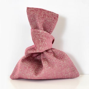 May include: A pink fabric gift bag with a knotted top. The bag is made of a textured fabric with a subtle white pattern.