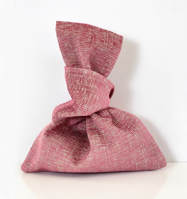 May include: A pink fabric gift bag with a knotted top. The bag is made of a textured fabric with a subtle white pattern.