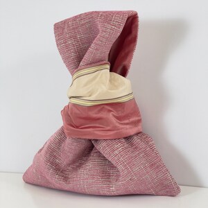 May include: A pink fabric bag with a knotted top. The bag is made of a textured fabric and has a pink satin lining with a beige and yellow striped trim.