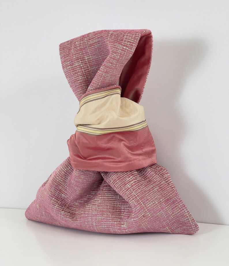 May include: A pink fabric bag with a knotted top. The bag is made of a textured fabric and has a pink satin lining with a beige and yellow striped trim.