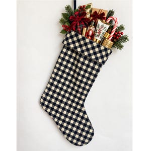 May include: A black and white buffalo check Christmas stocking filled with gifts including a nutcracker, a candy cane, small wrapped boxes with velvet bows, red berries, and evergreen sprigs. The stocking has a folded cuff at the top.