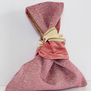 May include: A pink fabric bag with a knotted beige and pink ribbon. The bag is made of a textured fabric with a subtle sheen.