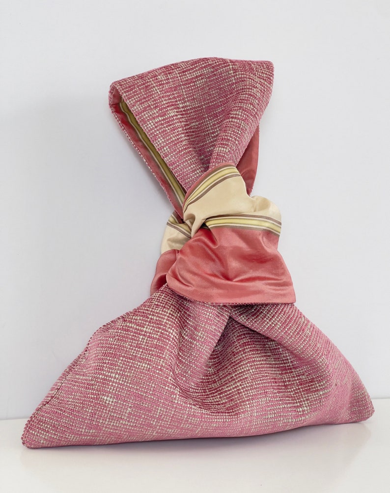 May include: A pink fabric bag with a knotted beige and pink ribbon. The bag is made of a textured fabric with a subtle sheen.