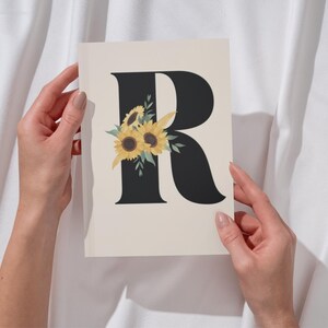 May include: A cream-colored card featuring a black letter "R" adorned with yellow sunflowers and green leaves. The card is held against a white fabric backdrop, creating a simple yet elegant design. The floral accent adds a touch of nature.