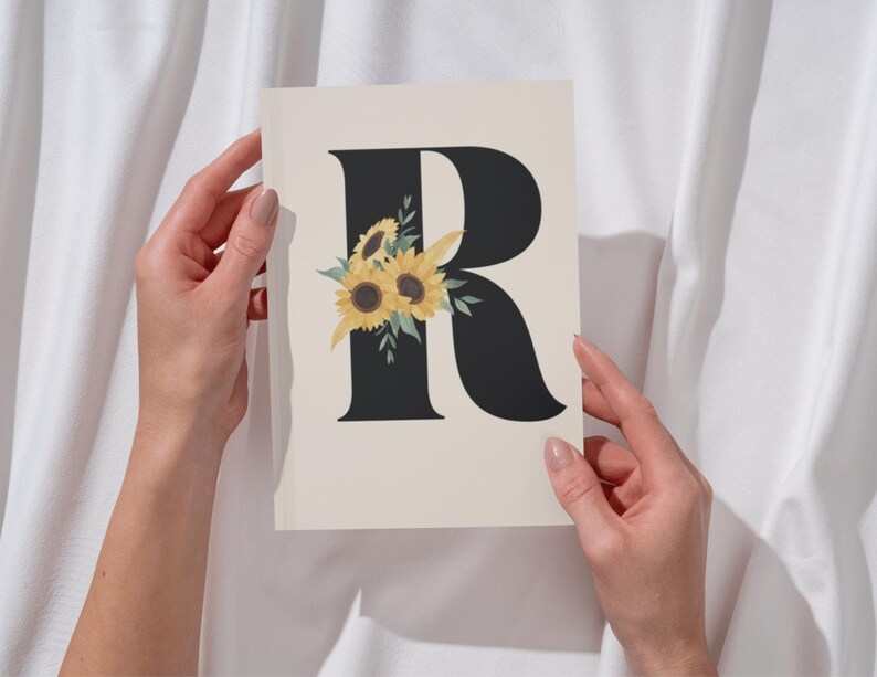 May include: A cream-colored card featuring a black letter "R" adorned with yellow sunflowers and green leaves. The card is held against a white fabric backdrop, creating a simple yet elegant design. The floral accent adds a touch of nature.
