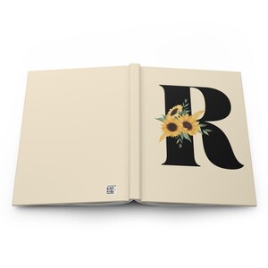 May include: Open journal with a cream-colored cover and pages. The right page features a large black letter "R" adorned with a cluster of sunflowers. The sunflowers have yellow petals and brown centers, with green leaves. The left page is blank.