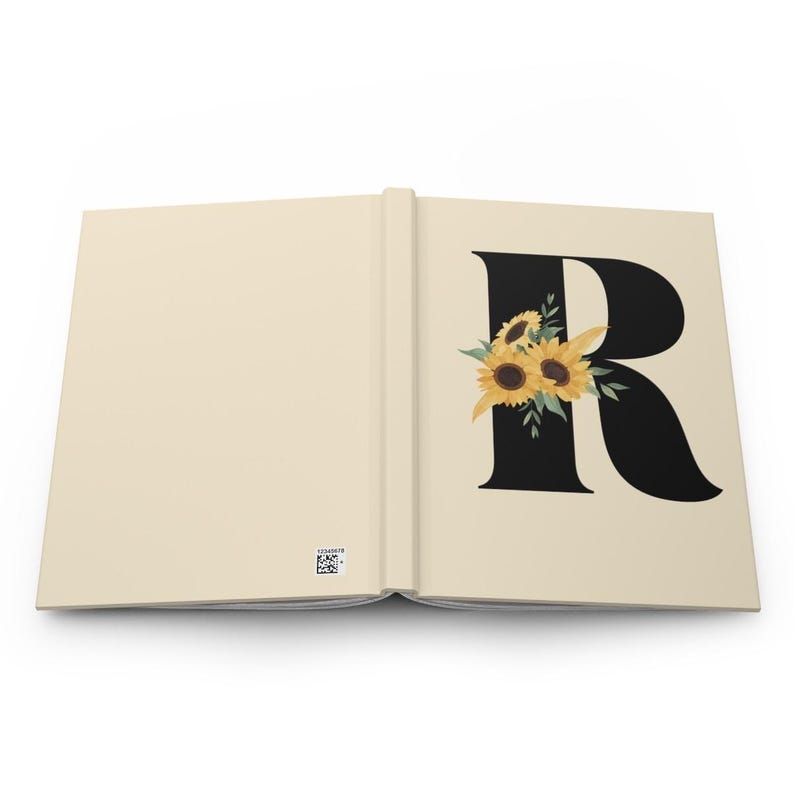 May include: Open journal with a cream-colored cover and pages. The right page features a large black letter "R" adorned with a cluster of sunflowers. The sunflowers have yellow petals and brown centers, with green leaves. The left page is blank.