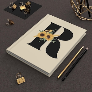 May include: A cream-colored journal featuring a large black letter "R" adorned with a sunflower bouquet. Gold and black pencils and gold binder clips are arranged around the journal on a dark surface.