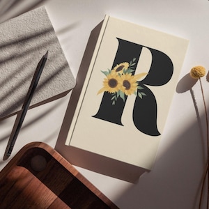 May include: A cream-colored journal with a large black letter "R" adorned with sunflower illustrations. A black pencil and a wooden tray are also visible, along with dried yellow flowers.
