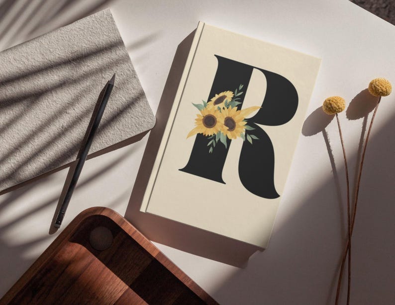 May include: A cream-colored journal with a large black letter "R" adorned with sunflower illustrations. A black pencil and a wooden tray are also visible, along with dried yellow flowers.
