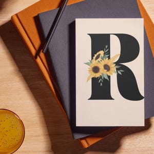 May include: A floral print card featuring a large black letter "R" adorned with sunflowers and greenery. The card is set against a neutral background, resting on a stack of notebooks and a pencil.