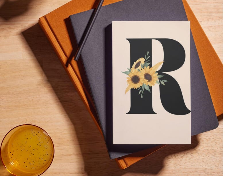 May include: A floral print card featuring a large black letter "R" adorned with sunflowers and greenery. The card is set against a neutral background, resting on a stack of notebooks and a pencil.